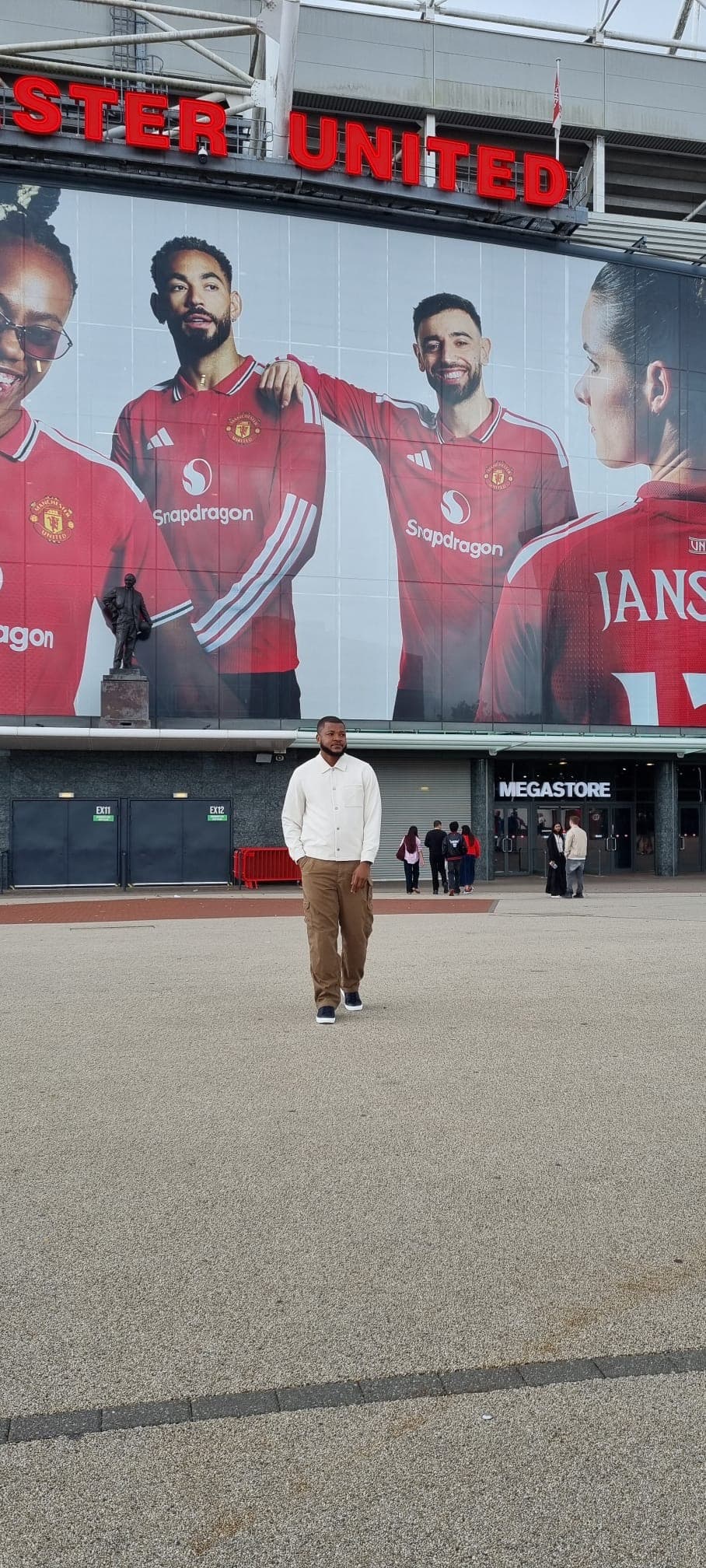 Dr Isaac Kwesi Acquah in a white shirt outside Old Trafford beneath a Manchester United player mural.